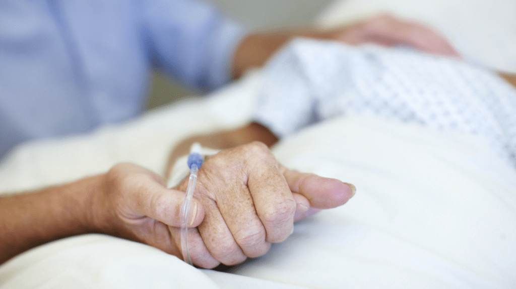 end of life care - person in hospital bed, IV in hand, holding the hand of someone else