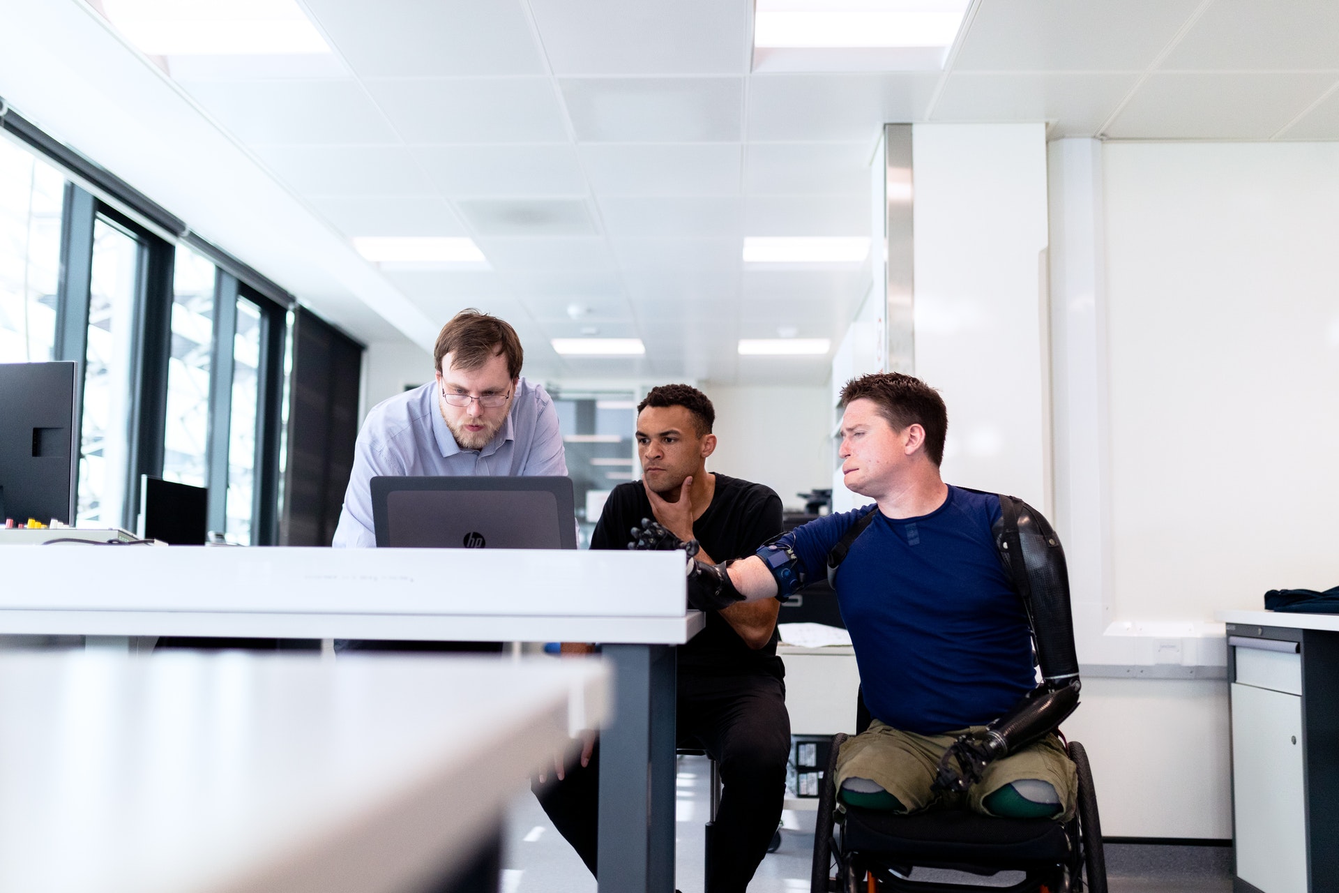 Three men looking at the computer and discussing. One man is disabled as he is on the wheel chair without legs and two bionic arms.