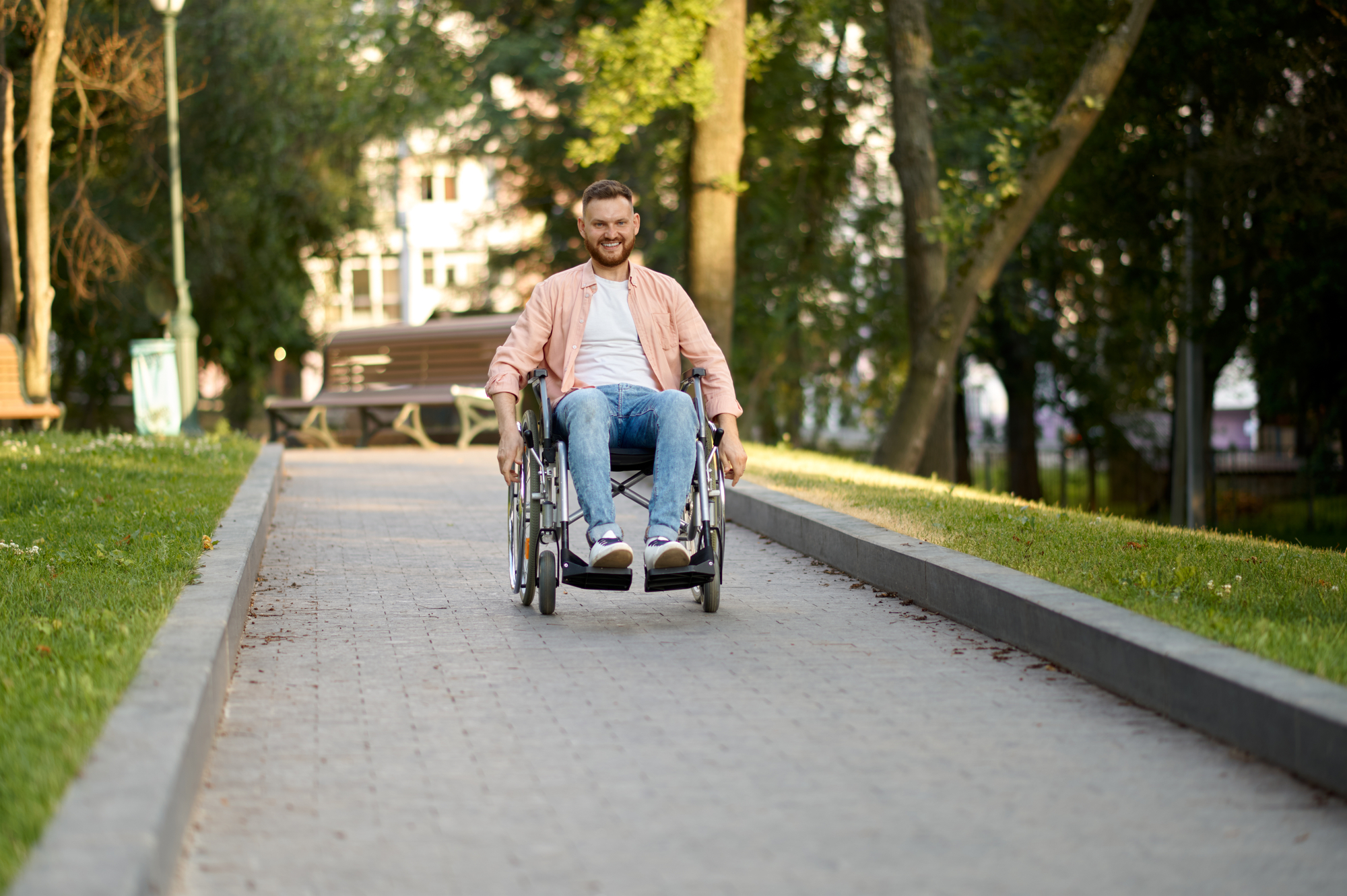 Disabled man in wheelchair rides on a path in the park. Paralyzed people and disability difficulties, handicap overcoming. Handicapped male person walking along the alley